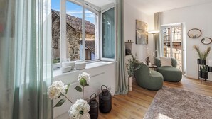 A modern living room with a large window, a green sofa, a wooden floor, and a view of a stone building outside. - Casa Oliva - Boutique Townhouse in Ascona (Ascona)
