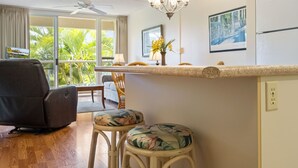 Una stanza con ventilatore a soffitto, un'area pranzo con tavolo e sedie, un bancone cucina con un vaso di fiori e vista sul verde dalla finestra.