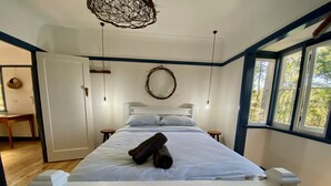 Une chambre avec un lit blanc, un plancher en bois, un luminaire suspendu et une fenêtre donnant sur des arbres.