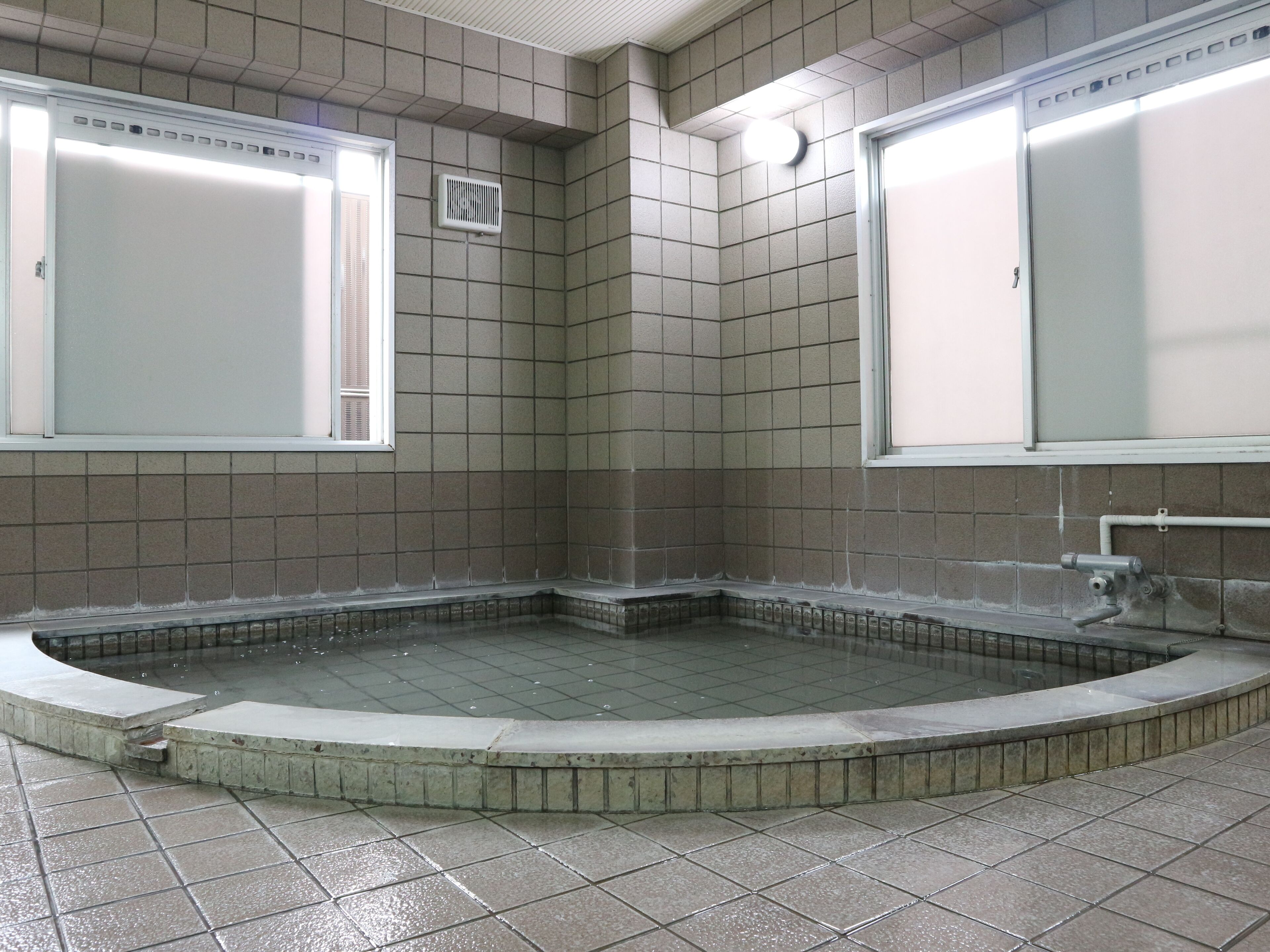 A public bath area with tiled walls and floor, a central water feature, and two large windows.