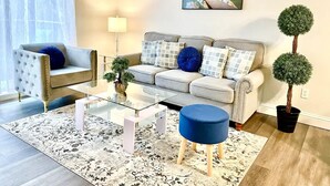A modern living room with a sofa, armchair, coffee table, and a small round ottoman. There is a potted plant and a painting on the wall.