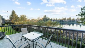 A balcony with a table and chairs overlooking a lake, surrounded by greenery and residential buildings.