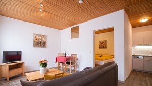 A modern living room with a grey sofa, wooden coffee table, and a TV stand. There is a dining area with a red tablecloth and chairs. A kitchen area is visible with white cabinets and a yellow cushion on the sofa. - Ferienwohnung Stettner See 42 qm (Rimsting)