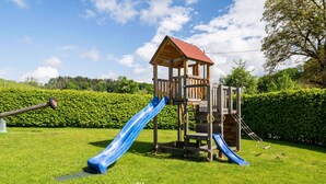 A wooden play structure with a red roof and two slides in a grassy area. - Ferienwohnung Stettner See 42 qm (Rimsting)