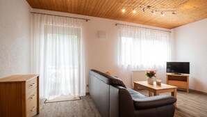 A modern living room with a sofa, a wooden table, a TV, and a window with sheer curtains. - Ferienwohnung Stettner See 42 qm (Rimsting)