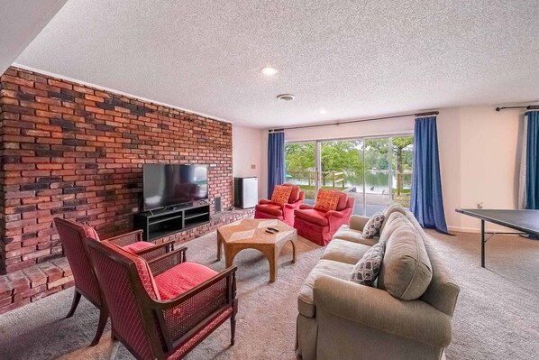 A living room with a brick wall, a flat-screen TV, red and beige furniture, and a view of a lake through large windows. - Great for Traveling Work Teams /20-30 minutes to downtown Atlanta/Sleeps 12/Private Pool (Jonesboro)