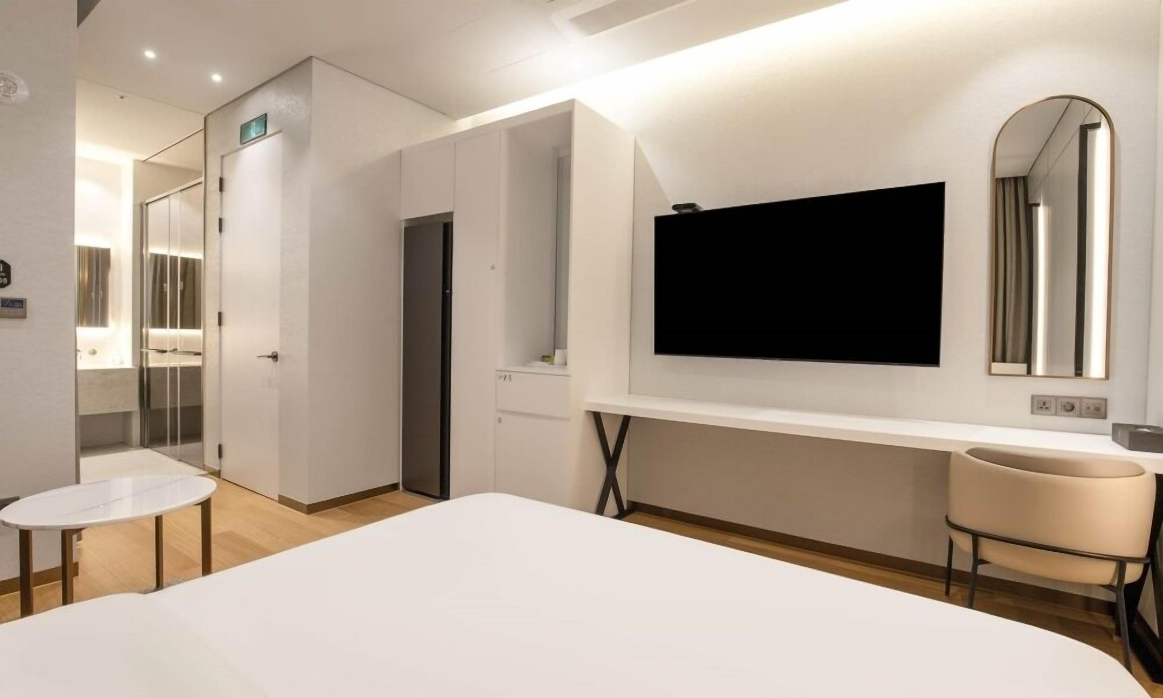 A modern hotel room with a large bed, a desk with a chair, a vanity with a mirror, and a television mounted on the wall.