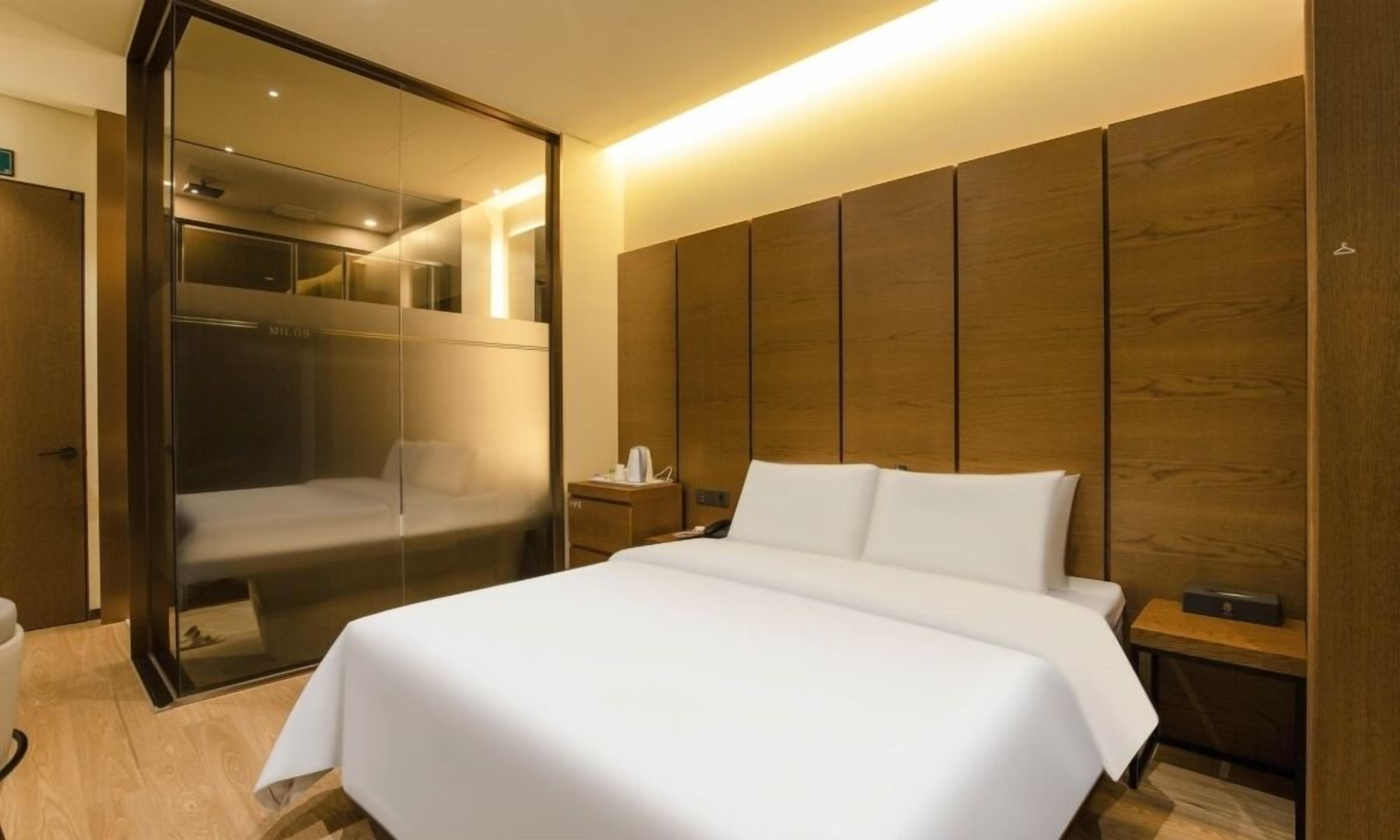 A hotel room with a large bed, a wooden headboard, a glass shower enclosure, and a bedside table with a lamp.