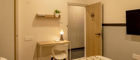 A hotel room with a bed, a desk with a chair, a small potted plant, a wall-mounted TV, and a door with a number.