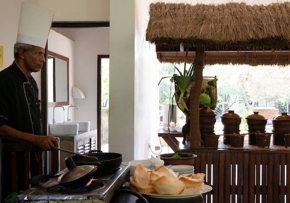 Un chef qui cuisine dans une cuisine dotée d’un toit de chaume, avec des casseroles et une vue sur l’extérieur.