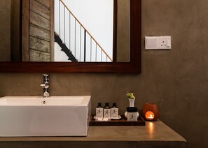 Une salle de bain avec un lavabo blanc, un miroir, un escalier en bois et un luminaire fixé au mur.