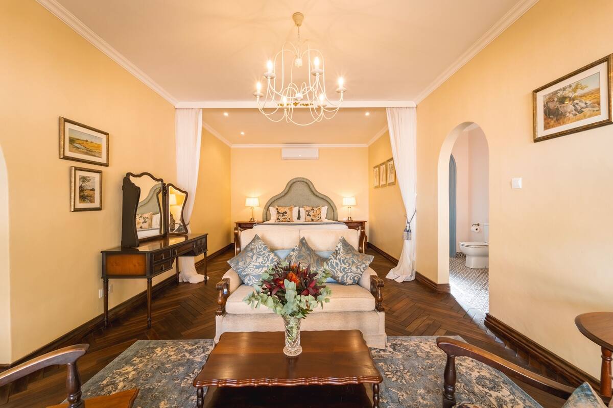 A hotel room with a large bed, a sitting area with a sofa and armchairs, a wooden coffee table with a vase of flowers, and a chandelier.