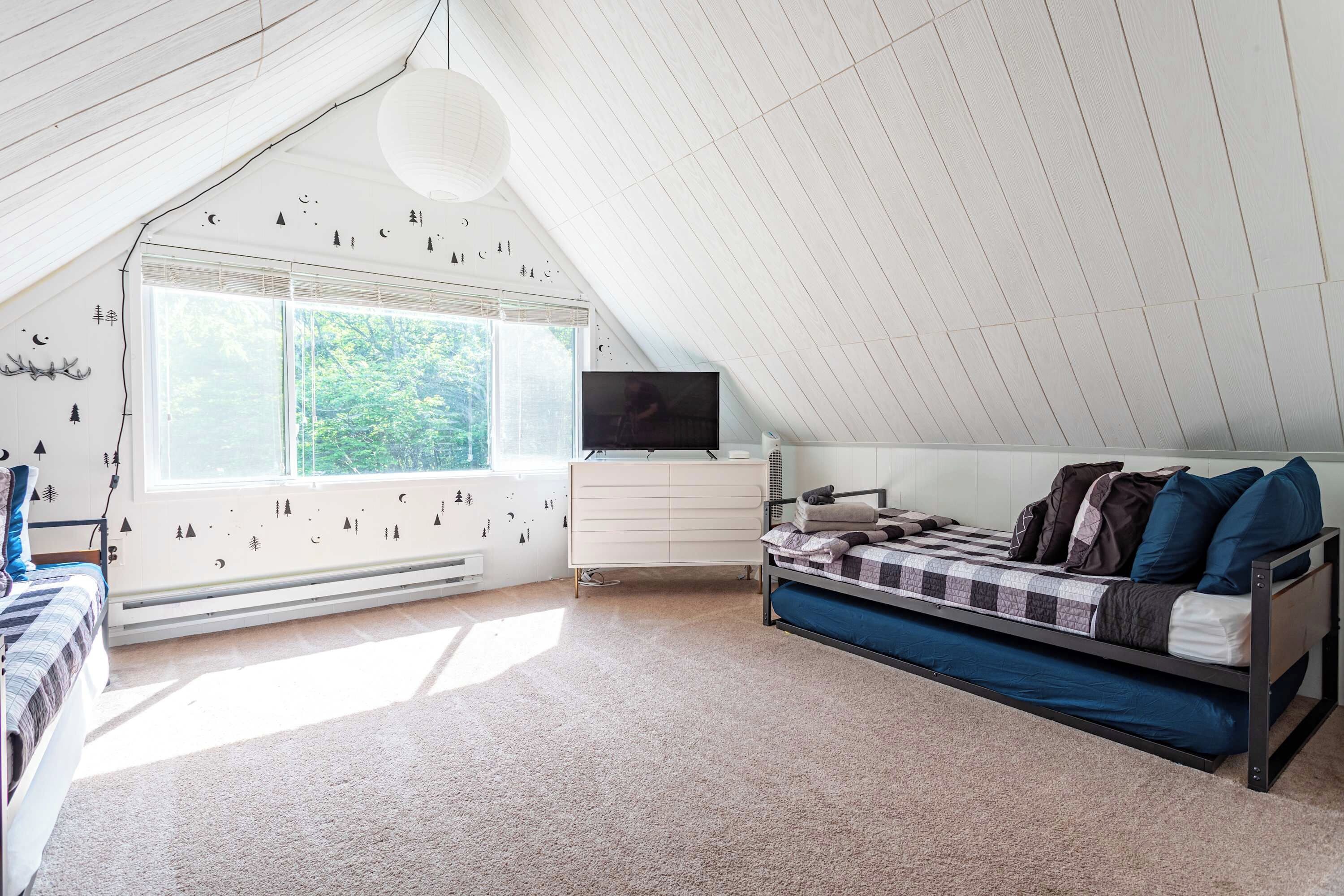 A bedroom with a sloped ceiling, a bed with a blue mattress, a dresser, a television, and a window with a view of trees.