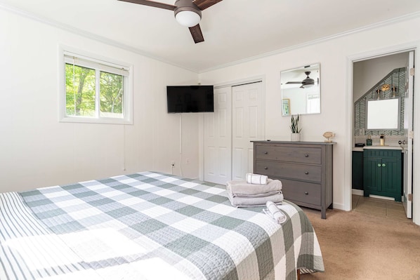 Un dormitorio con cama, cómoda, televisor, ventilador de techo y un baño visible a través de una puerta abierta.