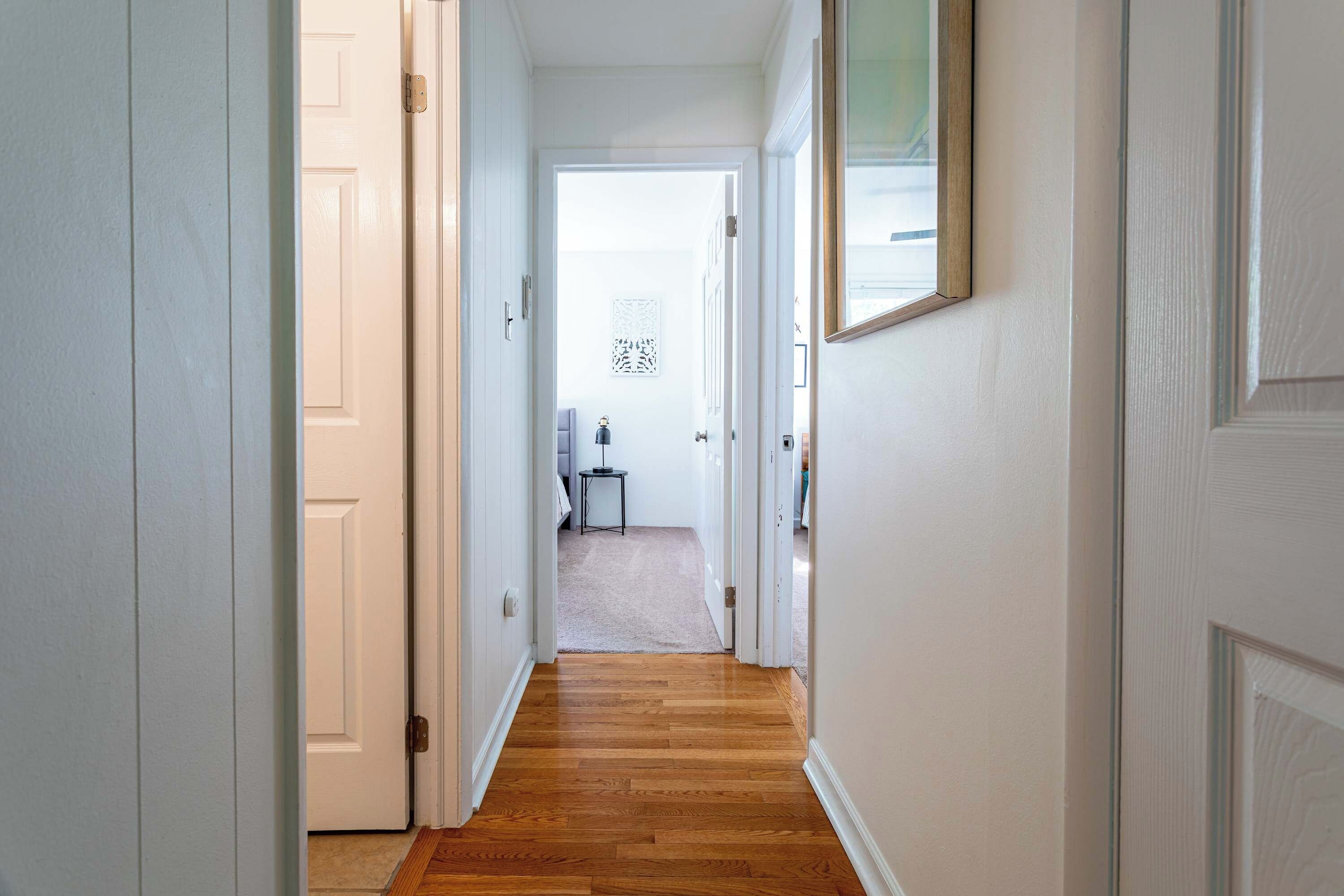 A hallway with white walls and wooden flooring leading to a room with a bed and a small table.