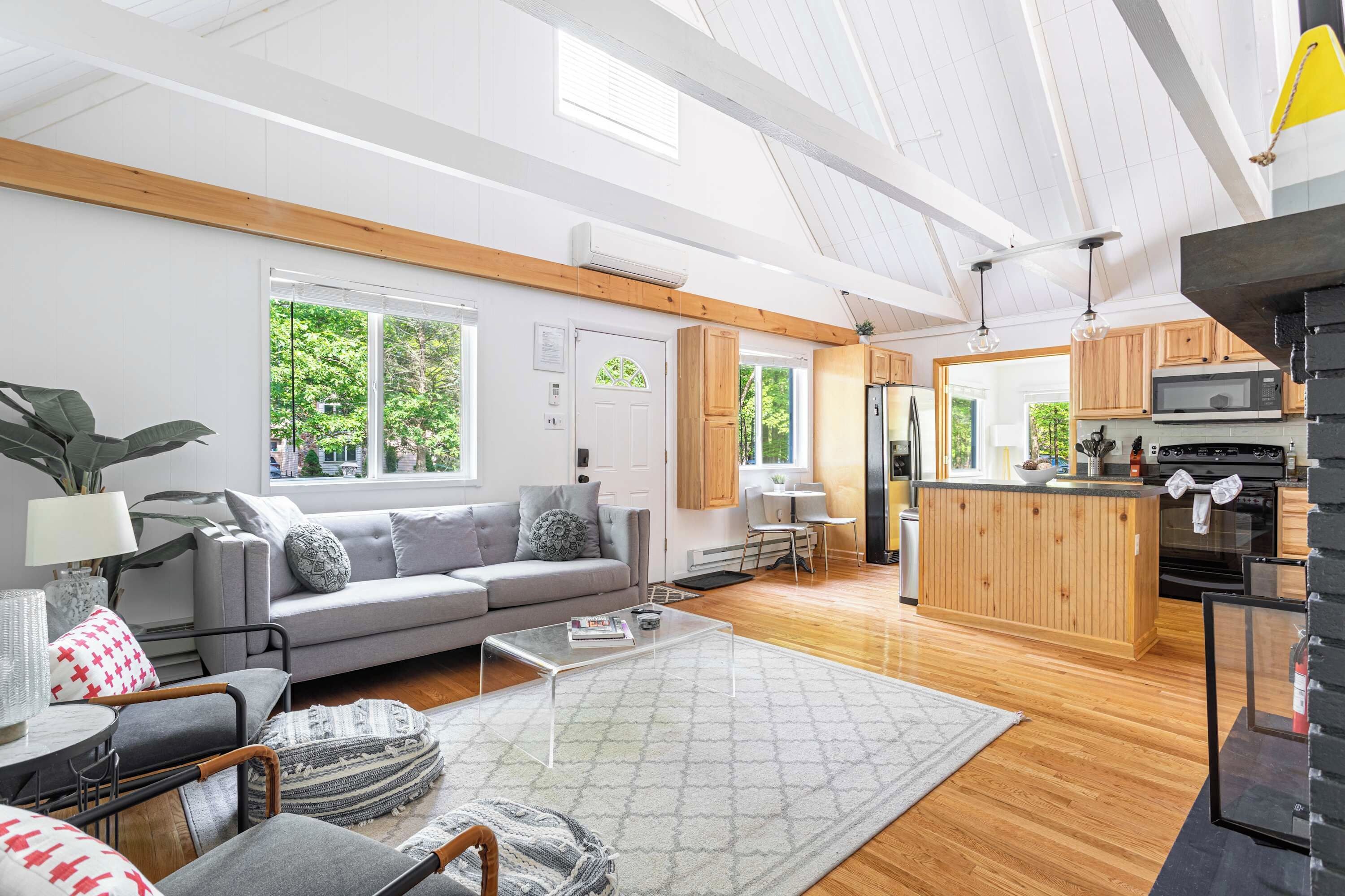 A modern living room with a grey sofa, wooden beams, a kitchen with a wooden island, and large windows.