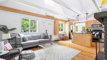 A modern living room with a grey sofa, wooden beams, a kitchen with a wooden island, and large windows.