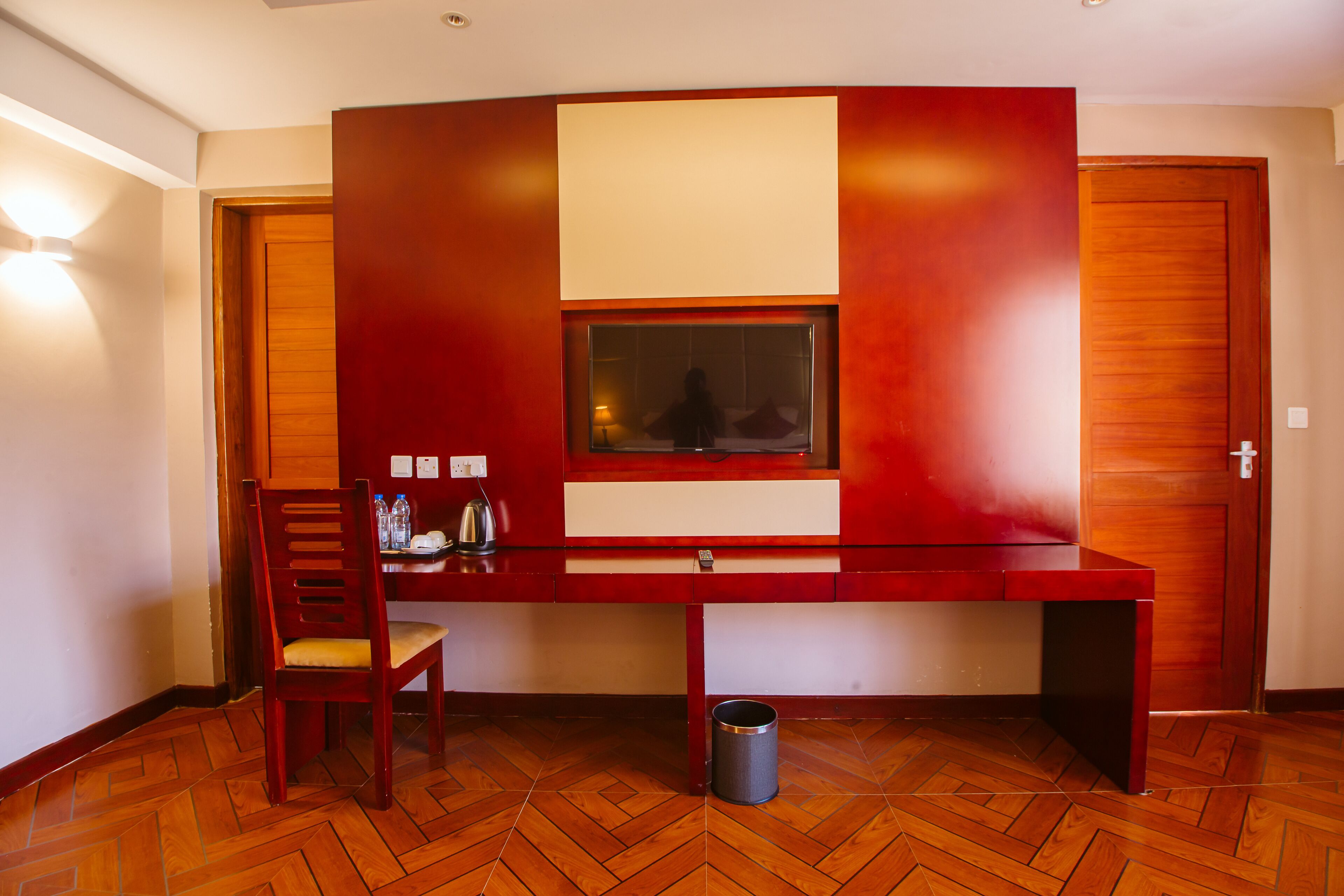 A modern room with a wooden herringbone floor, a red accent wall, a large window, a desk, a chair, and a trash can.