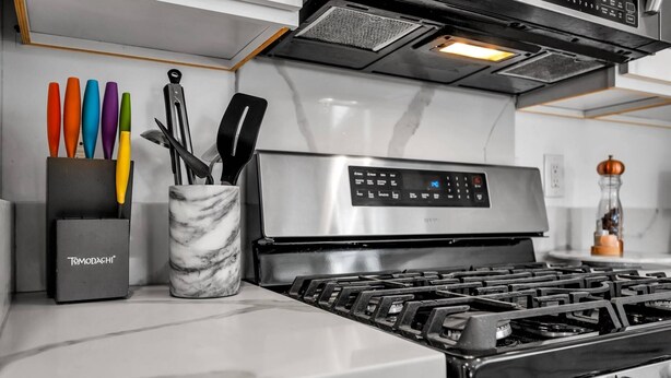 Una cocina con estufa de acero inoxidable, un recipiente de mármol que contiene utensilios de cocina, y una caja negra con la etiqueta 'Tomodachi'.