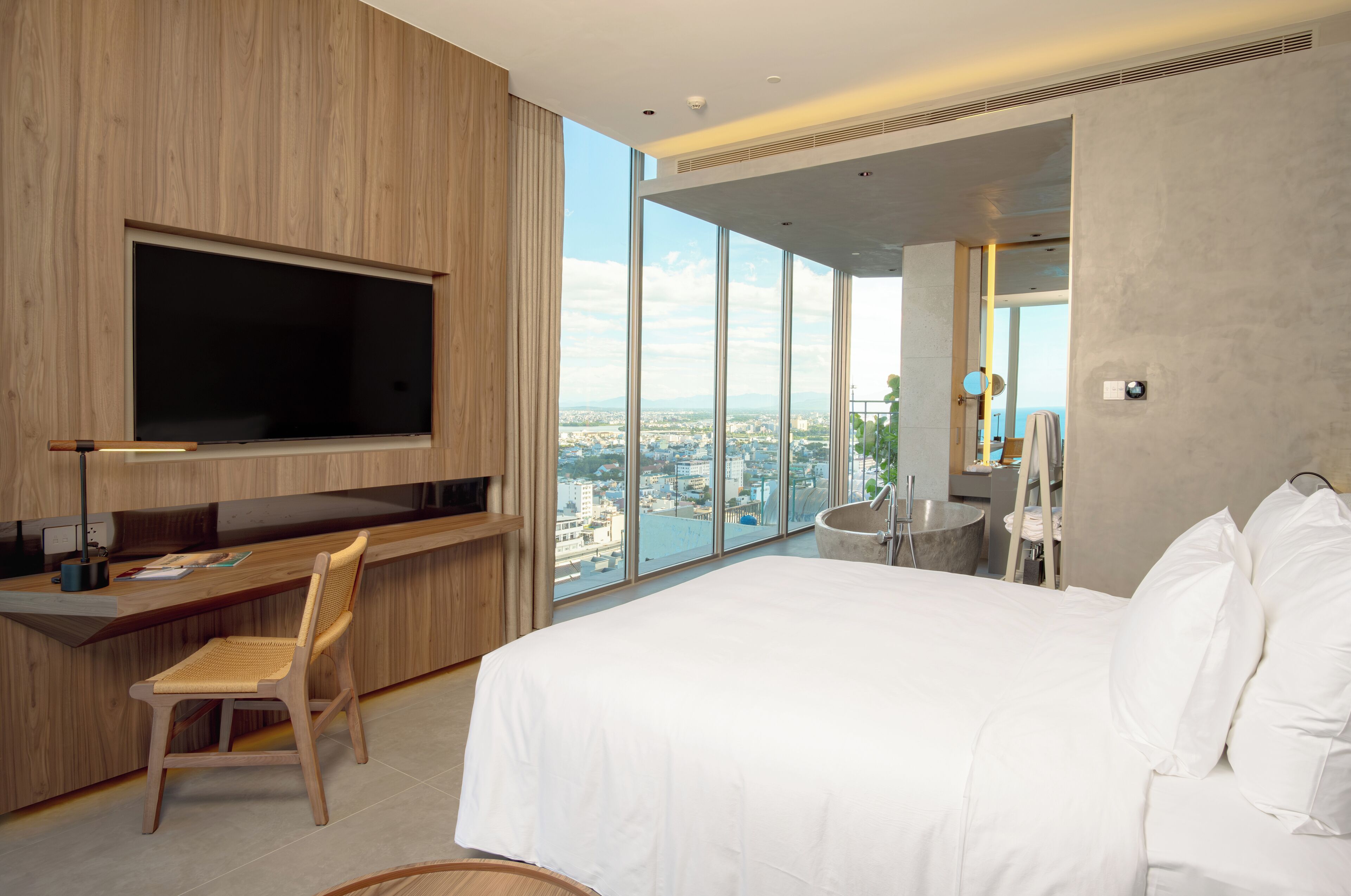 Une chambre d’hôtel moderne dotée d’un grand lit, d’un bureau avec une chaise, d’une télévision et offrant une vue sur la ville grâce à de grandes fenêtres.
