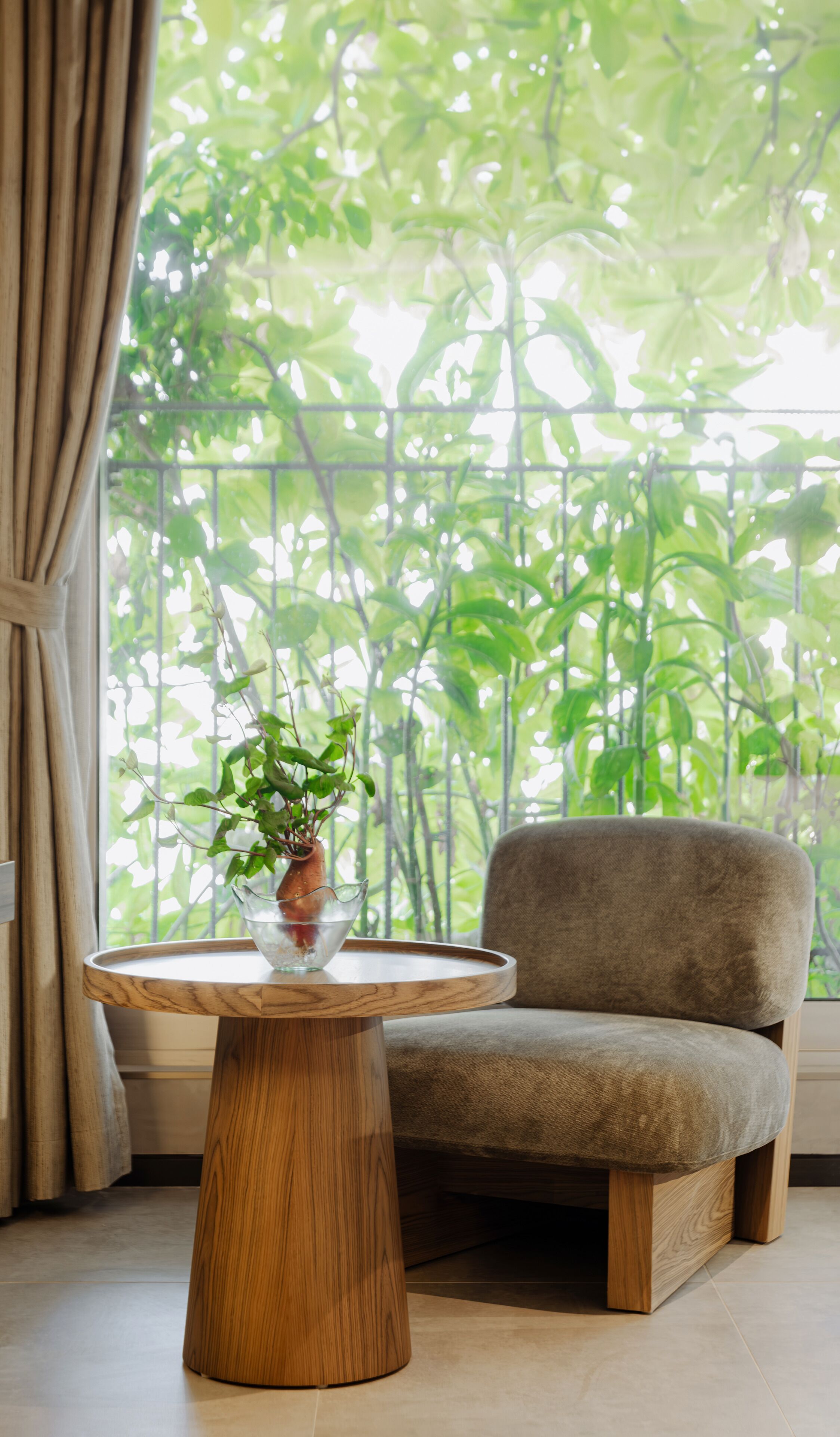 A room with a round wooden table, a potted plant, and a grey armchair by a window with a view of green foliage.