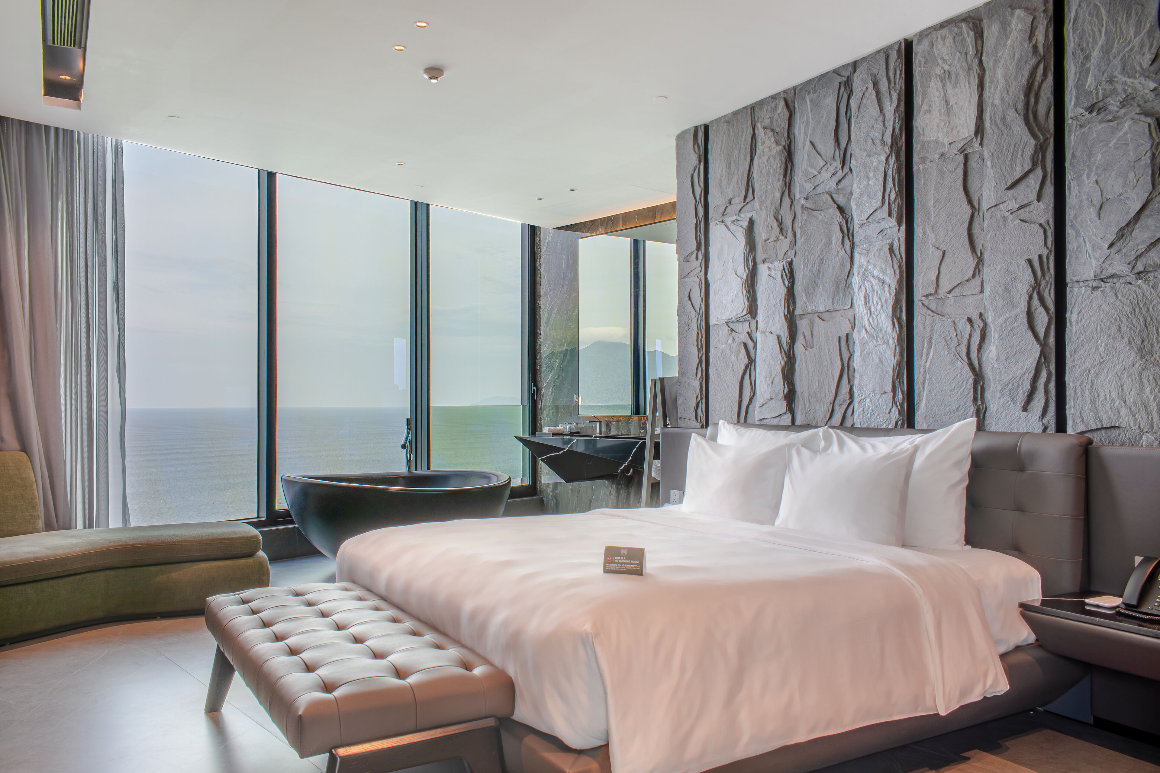 A modern hotel room with a large bed, a seating area, and a view of the sea.