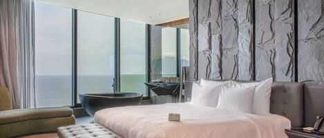 A modern hotel room with a large bed, a seating area, and a view of the sea.
