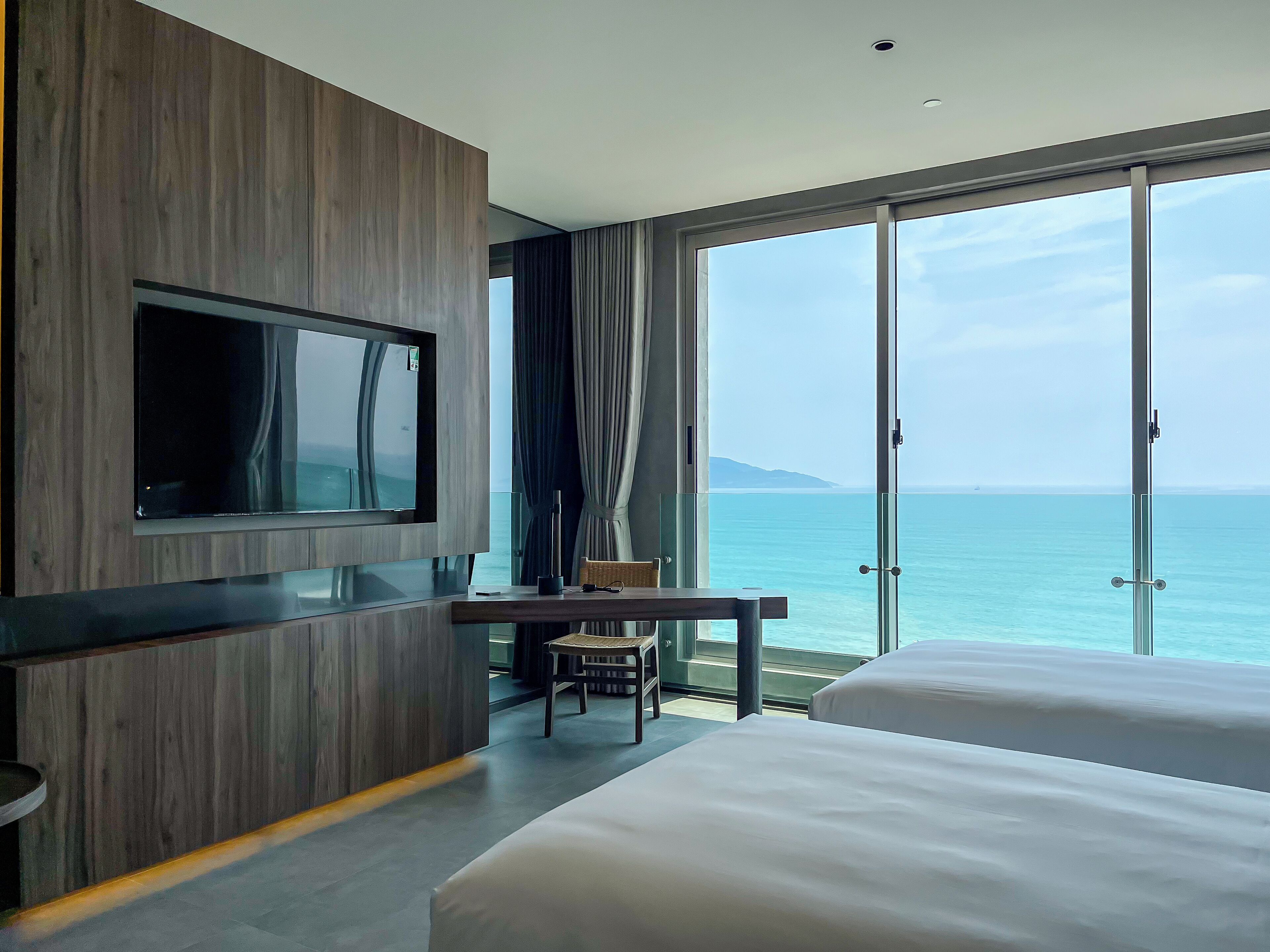 Quarto de hotel moderno com uma televisão grande, uma escrivaninha e vista para o mar.
