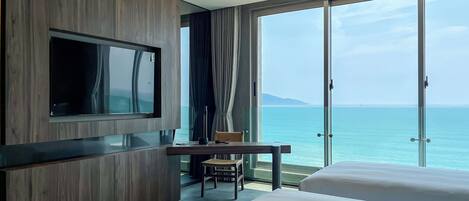 Quarto de hotel moderno com uma televisão grande, uma escrivaninha e vista para o mar.