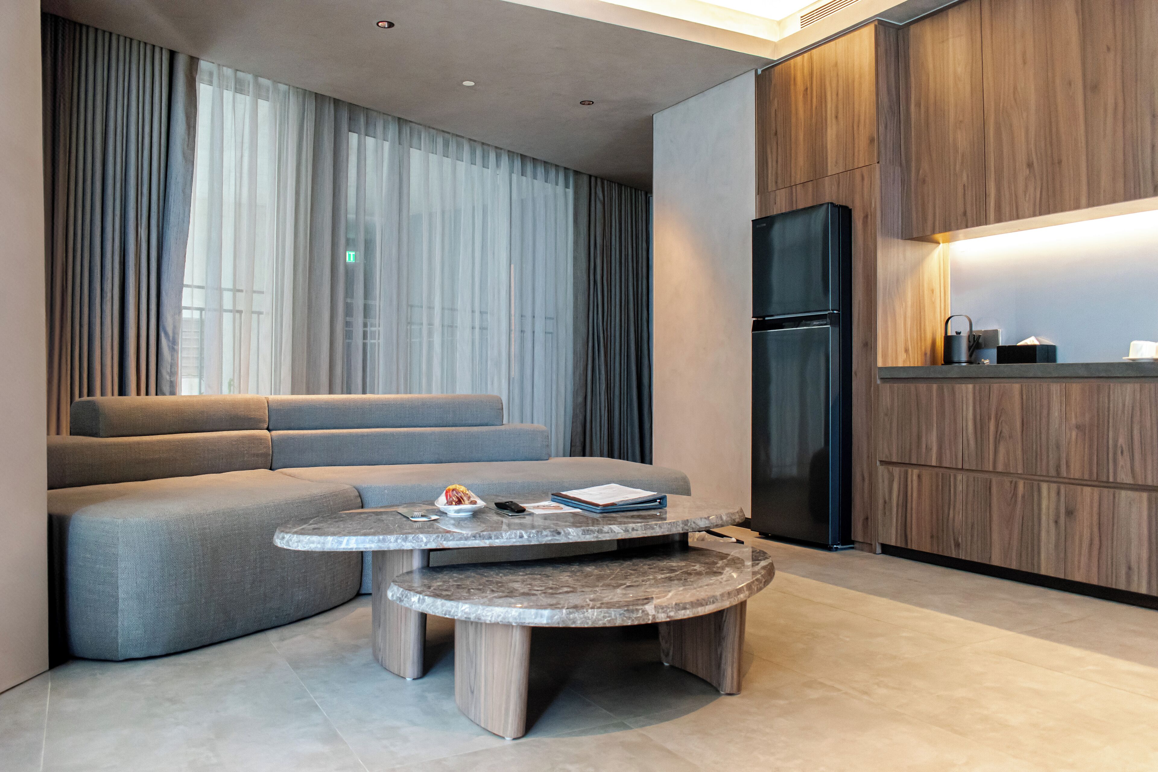 Un salón moderno con un sofá gris, una mesa de centro redonda y una cocina con armarios de madera y un refrigerador negro.