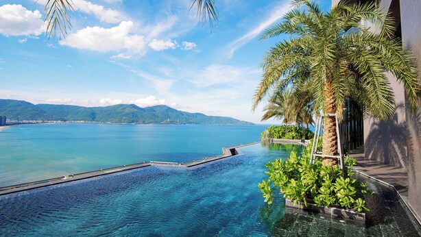 A pool with a view of the sea and mountains, surrounded by palm trees and modern architecture. - M HOTEL DANANG (Da Nang)