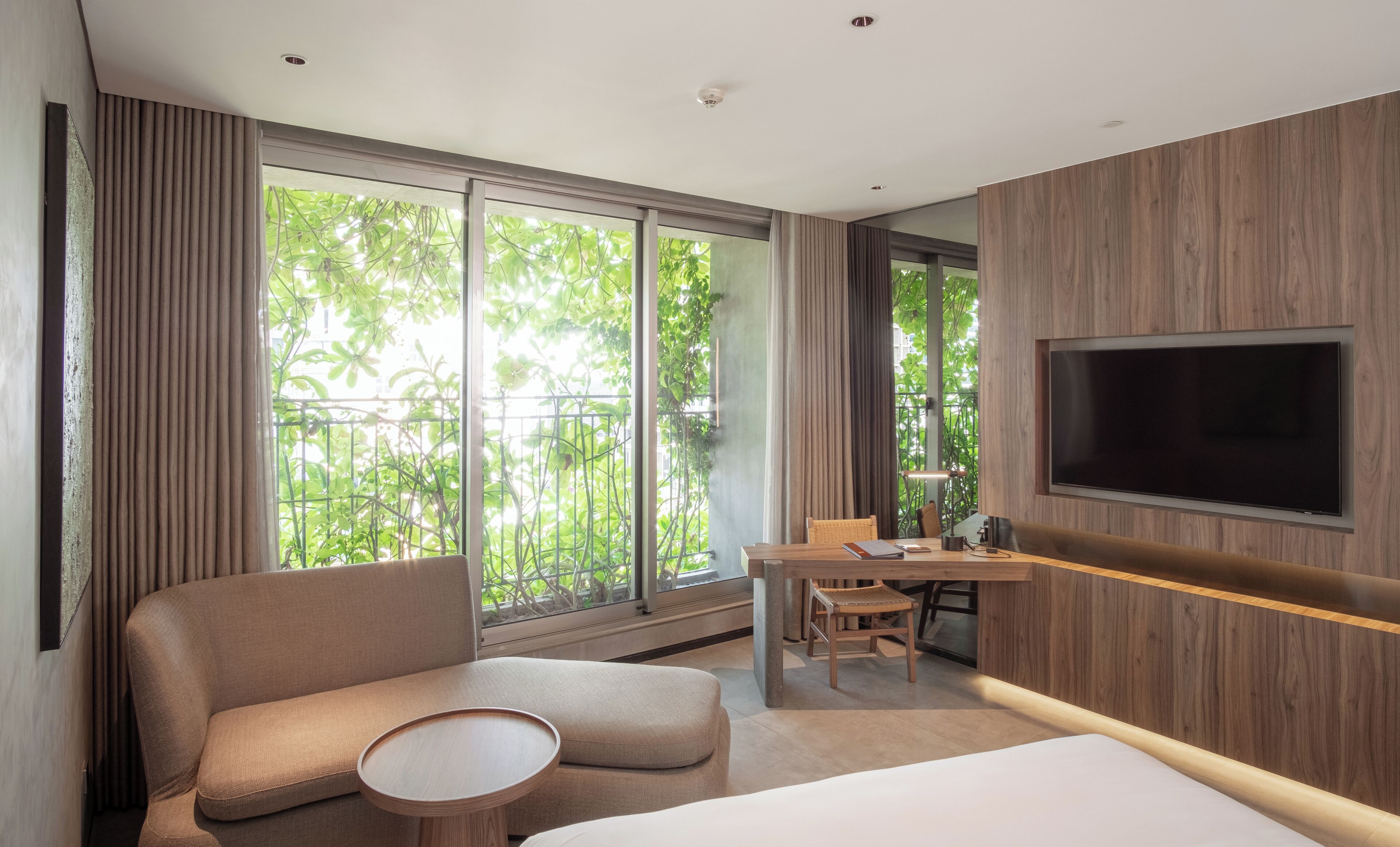 Een moderne hotelkamer met een groot raam, een bank, een klein rond tafeltje, een bureau met stoel en een wandgemonteerde flatscreen televisie.