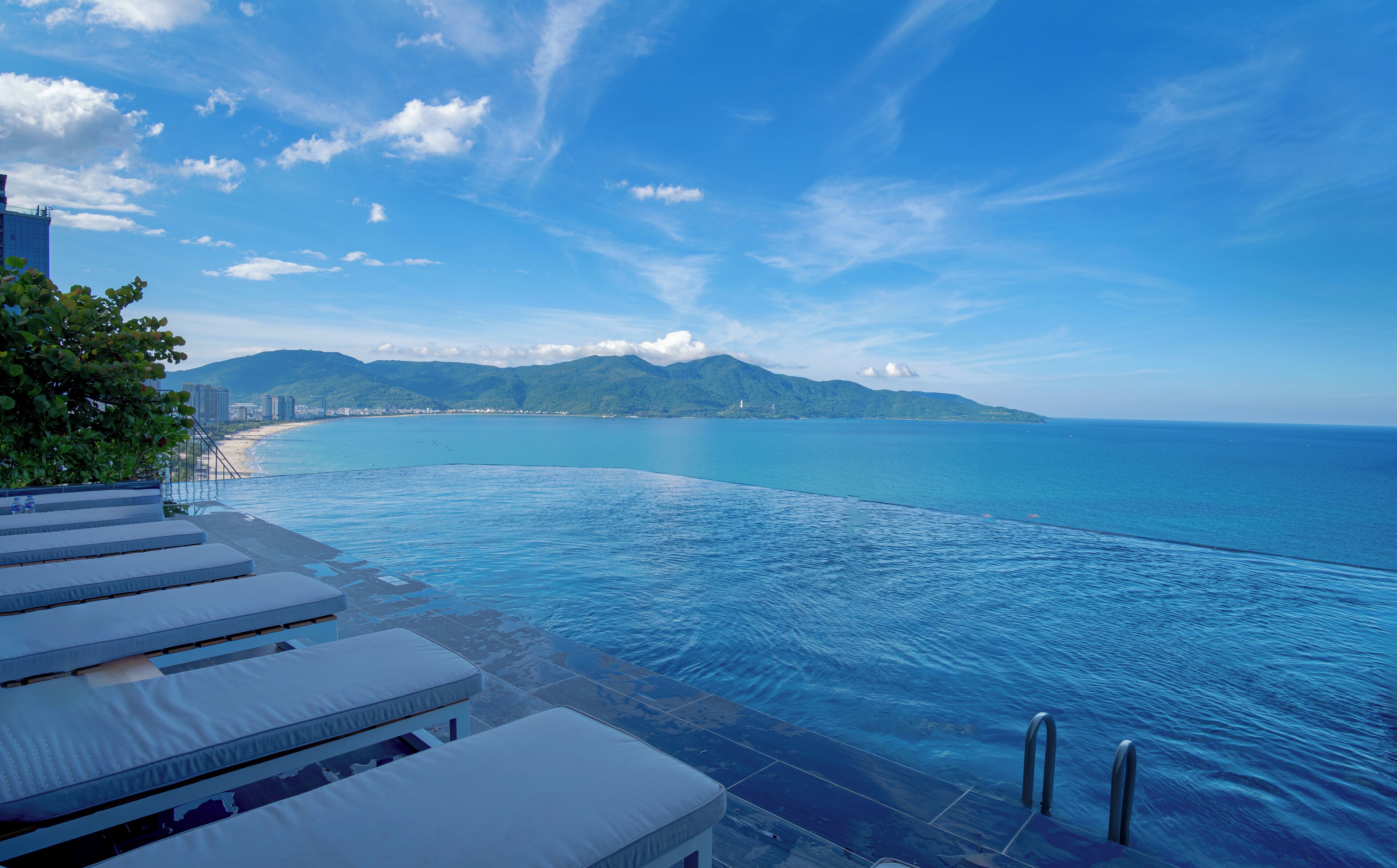 An infinity pool with a view of the sea and mountains, lounge chairs, and a clear sky.
