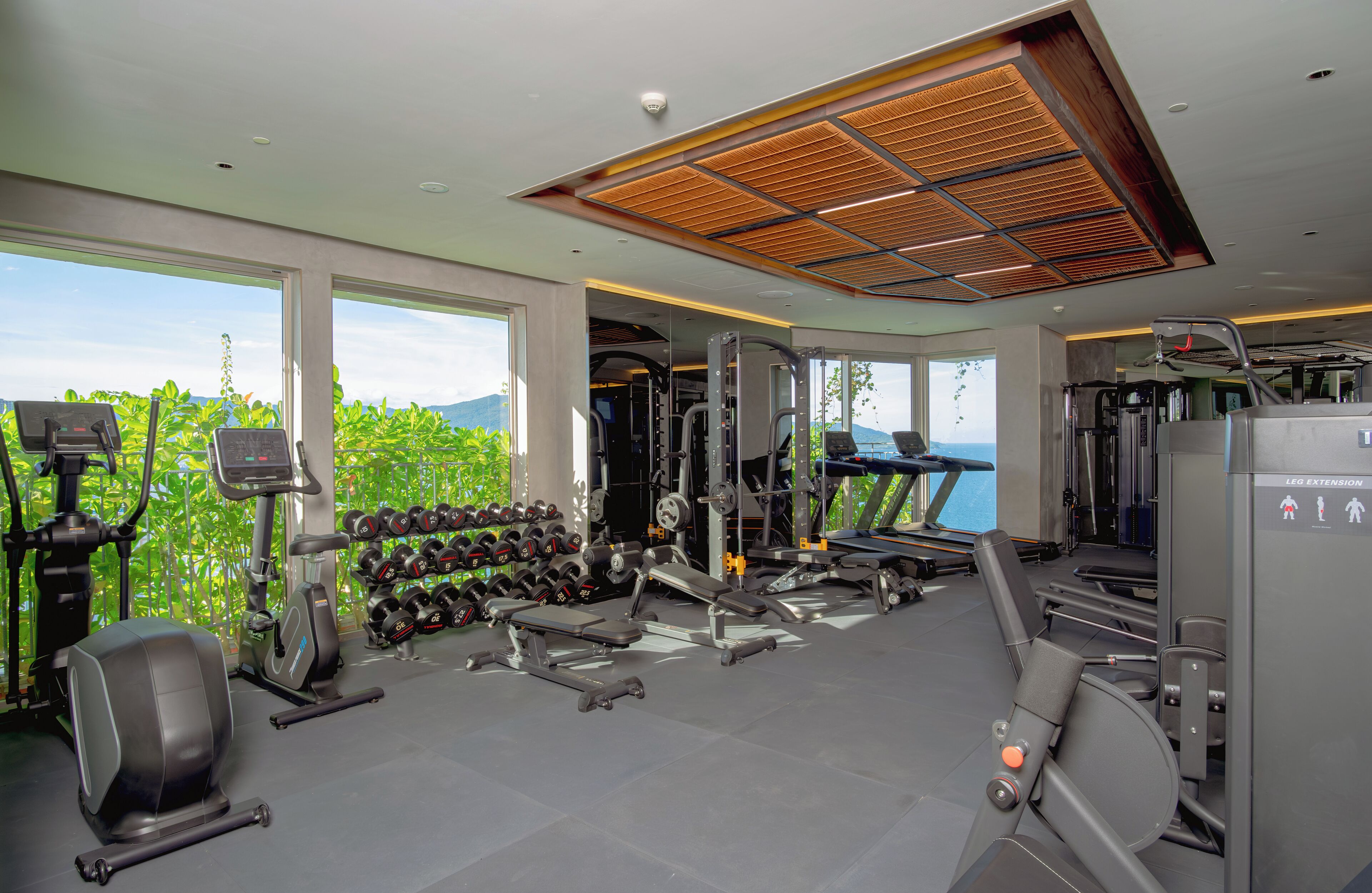 A gym with various exercise equipment, large windows with a view of greenery and mountains, and a ceiling with a wooden panel design.
