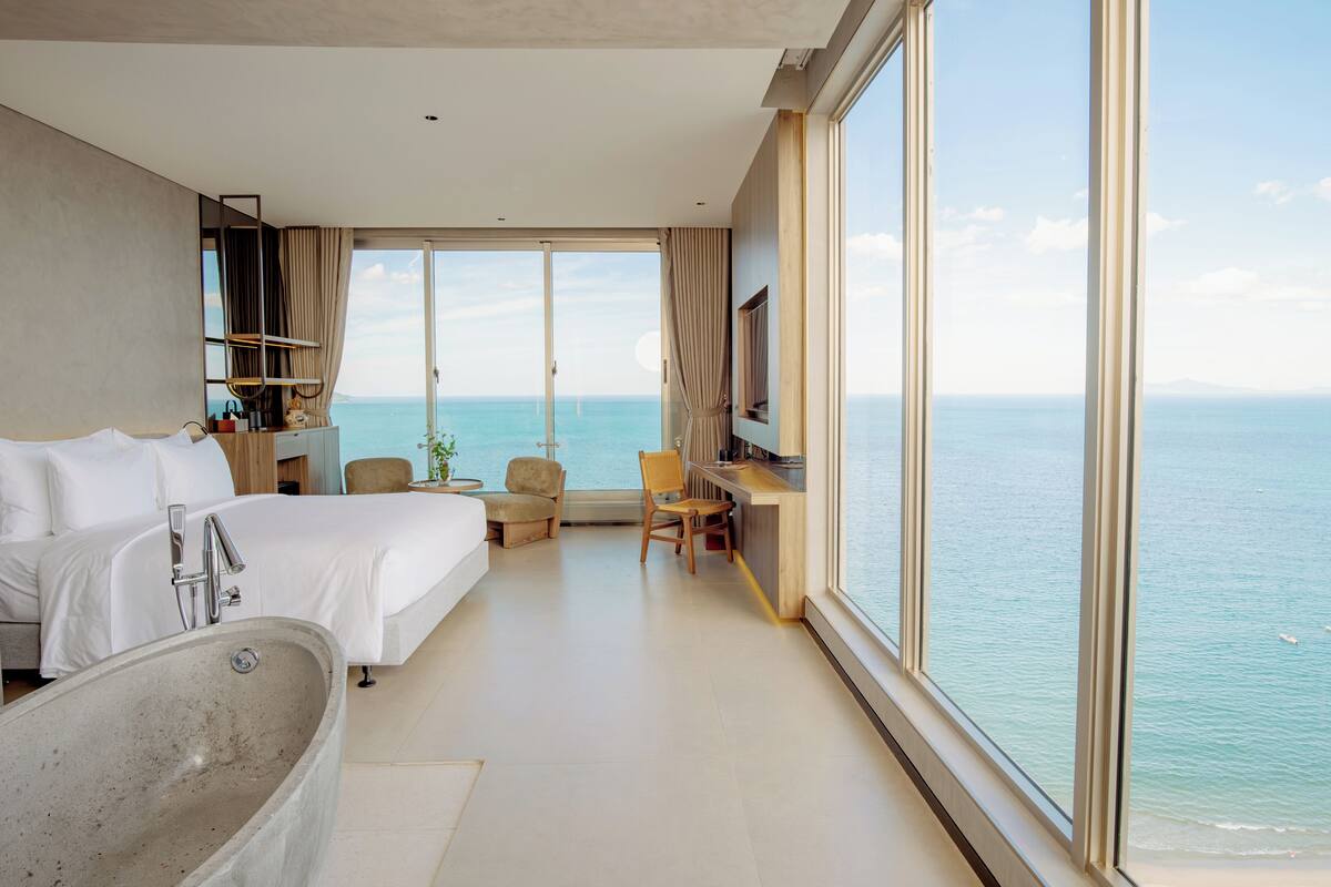 A modern hotel room with a large bed, a bathtub, a desk, and a view of the sea.
