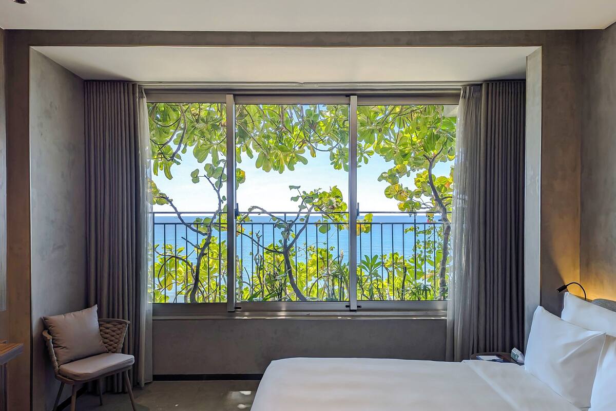 A hotel room with a large window, a bed, a chair, and a view of the sea.