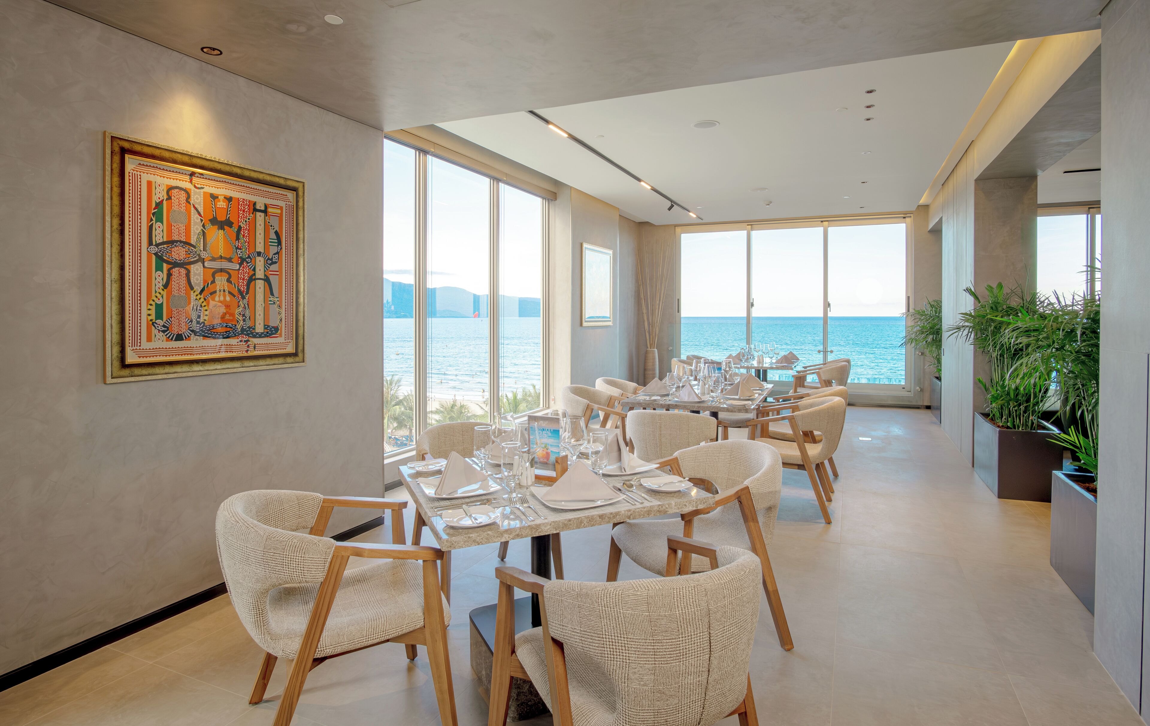 A dining area with tables set for guests, large windows offering a view of the sea, and a framed artwork on the wall.