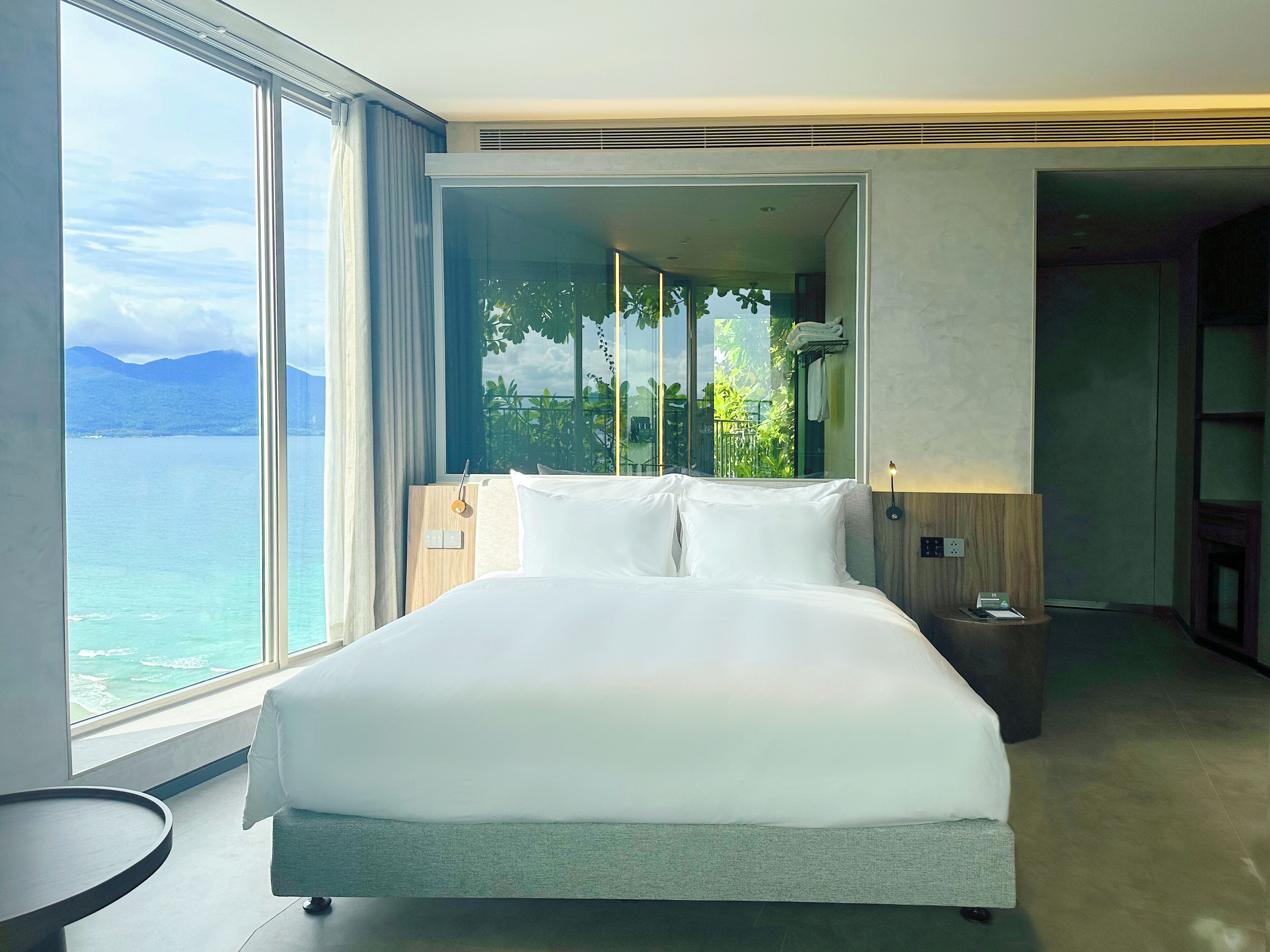 A hotel room with a large bed, a bedside table, a nightstand, and a view of the sea.