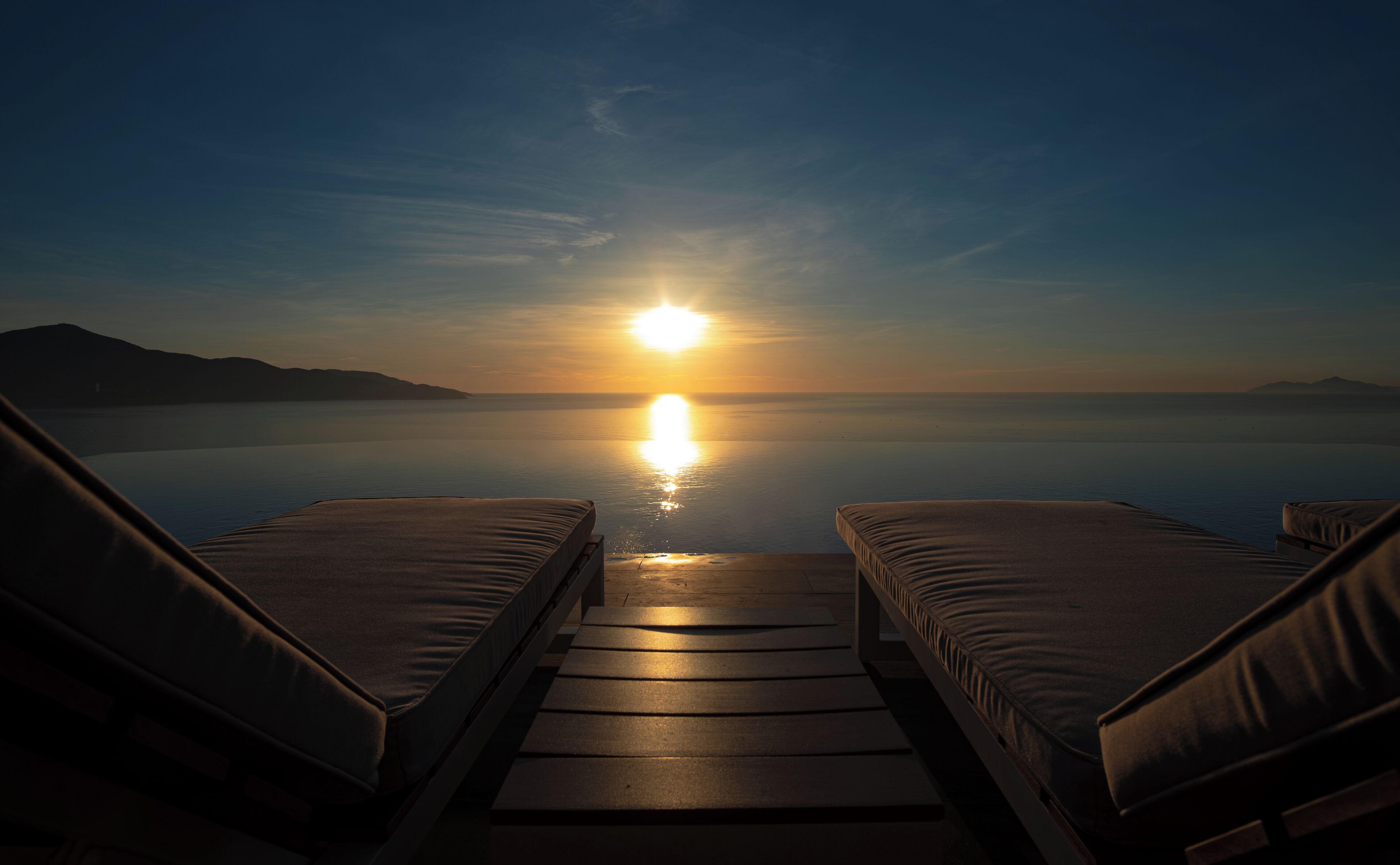 A sunset view from a lounge area with two sun loungers facing a calm sea and distant mountains.