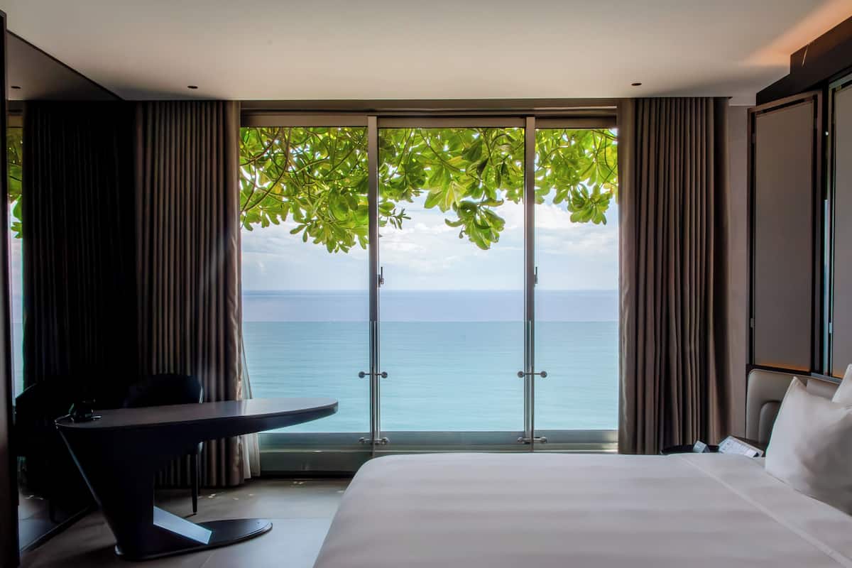 A hotel room with a large bed, a desk, and a view of the sea through floor-to-ceiling windows.