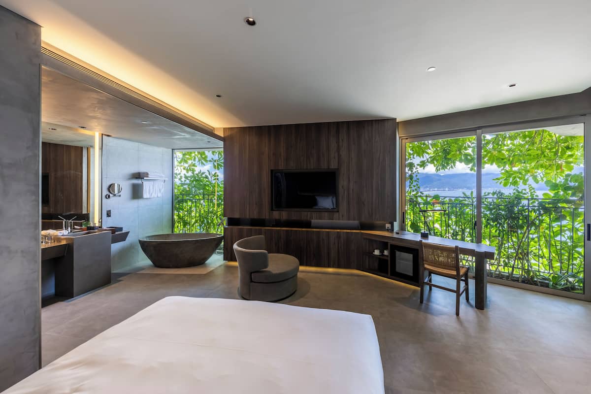 A modern hotel room with a large bed, a sitting area with a round chair, a large bathtub, a TV mounted on the wall, and a balcony with a view of greenery and mountains.