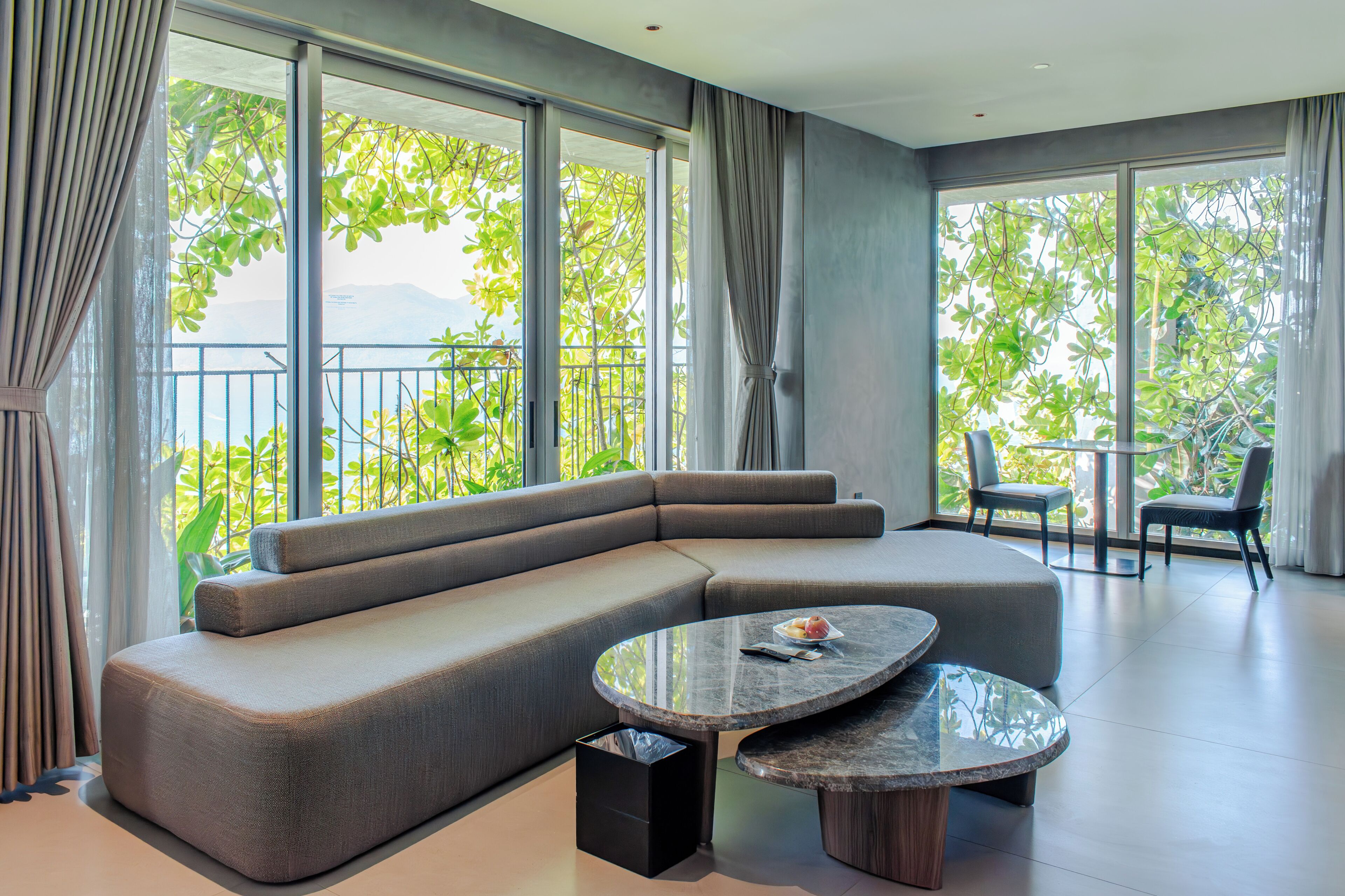 A modern living room with a sectional sofa, a round marble coffee table, and large windows with a view of greenery.