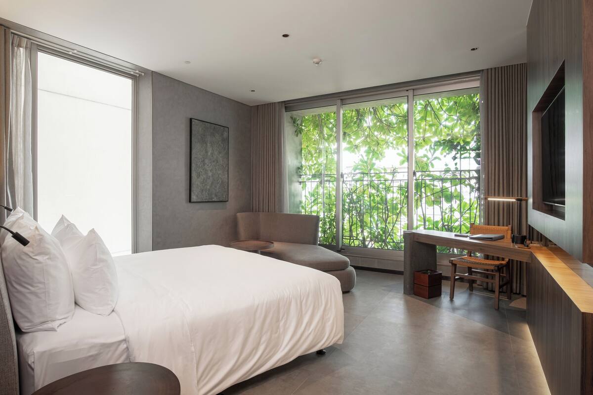 A modern hotel room with a large bed, a desk, and a view of greenery through the window.