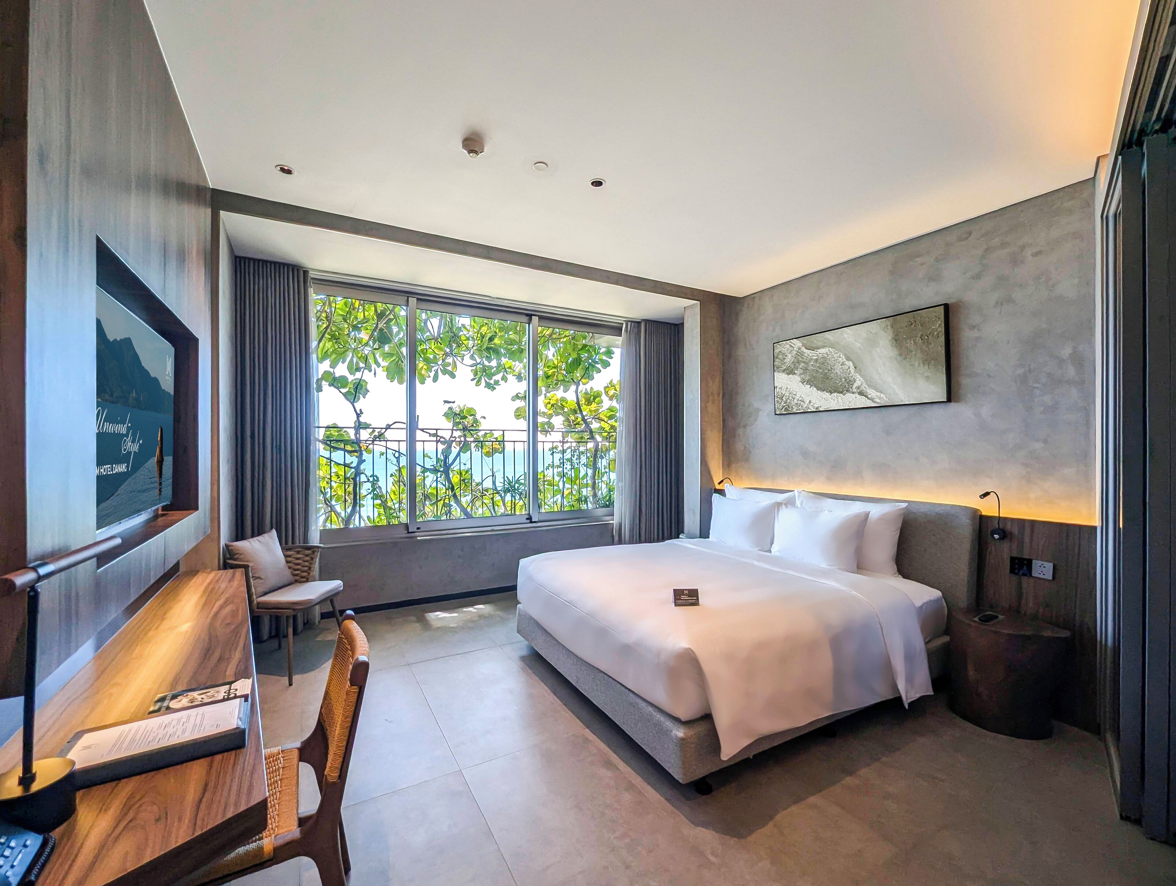 Habitación de hotel moderna con una cama grande, un escritorio y vistas a zonas verdes y montañas a través del ventanal.