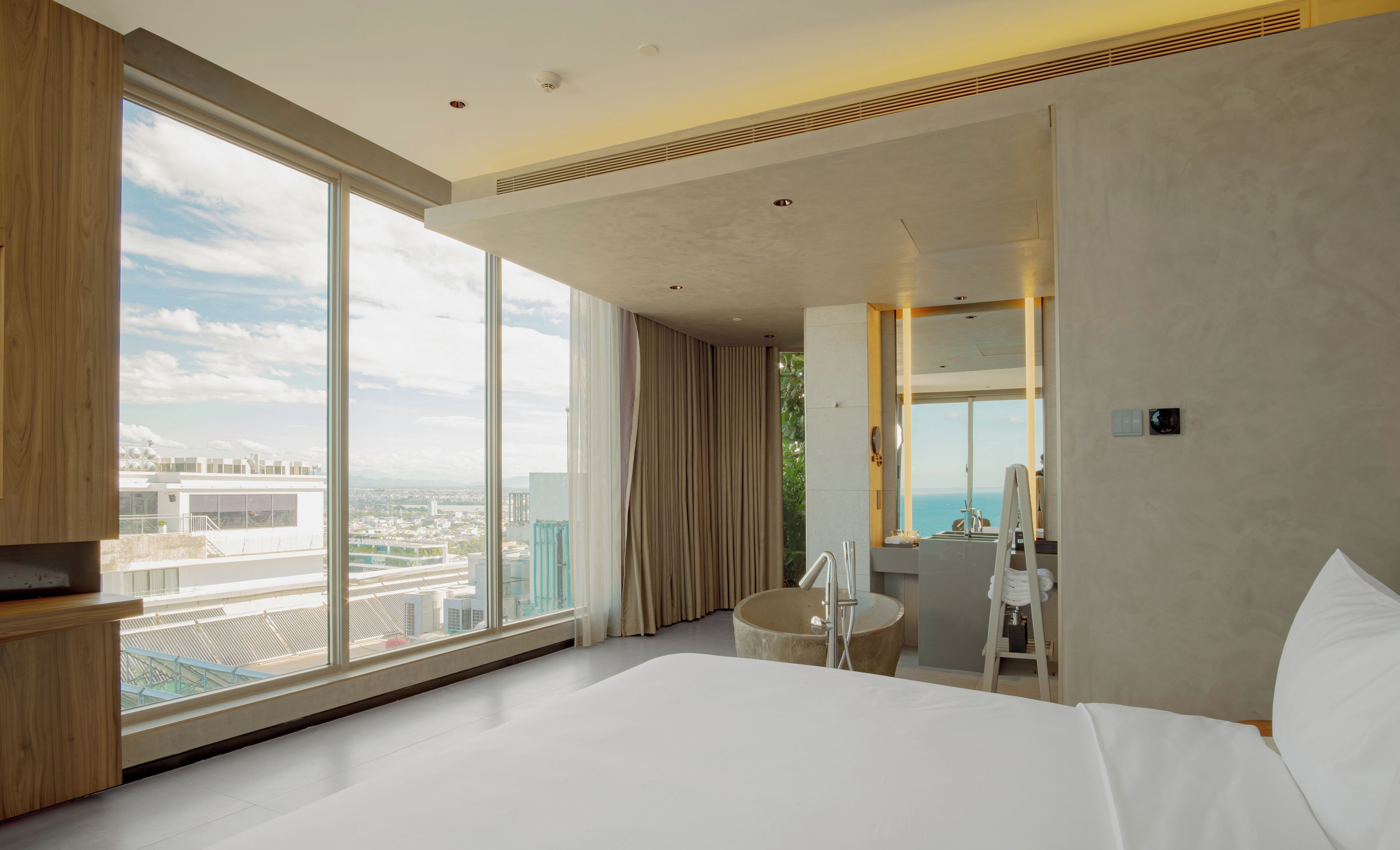 Una habitación de hotel moderna con una cama grande, un baño con bañera y vistas a la ciudad a través de amplios ventanales.