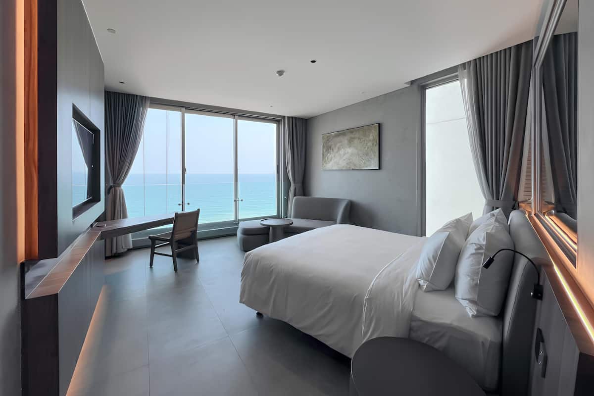 A modern hotel room with a large bed, a desk, and a chair. There is a large window with a view of the sea.