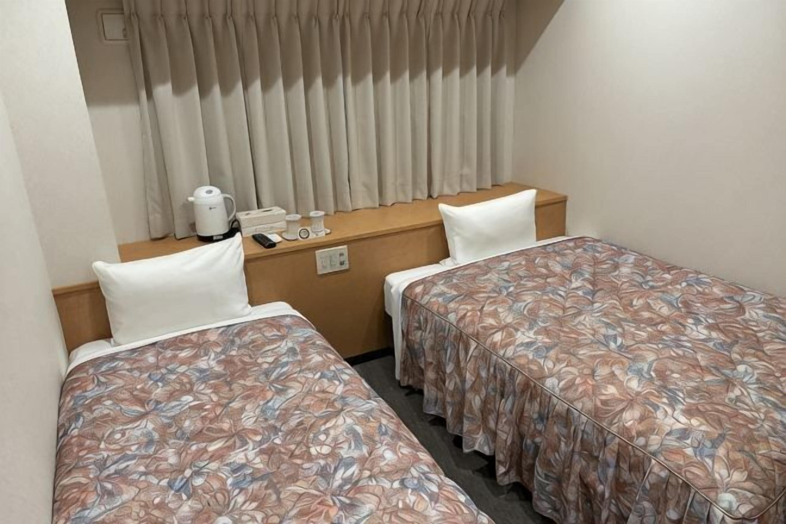 Une chambre d’hôtel avec deux lits simples, une table de chevet en bois avec une bouilloire, un téléphone et une fenêtre avec des rideaux.