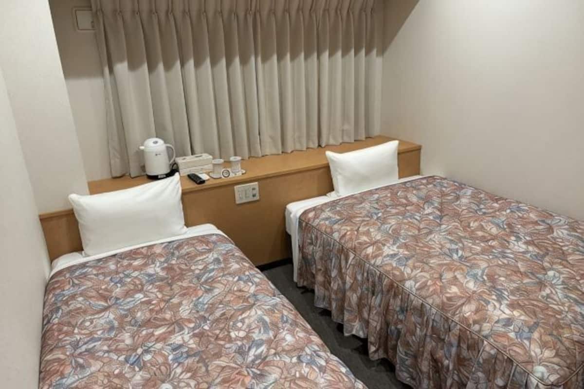 Ett hotellrum med tvÄ separata sÀngar, ett trÀbord vid sÀngen med en vattenkokare, en telefon och ett fönster med gardiner.