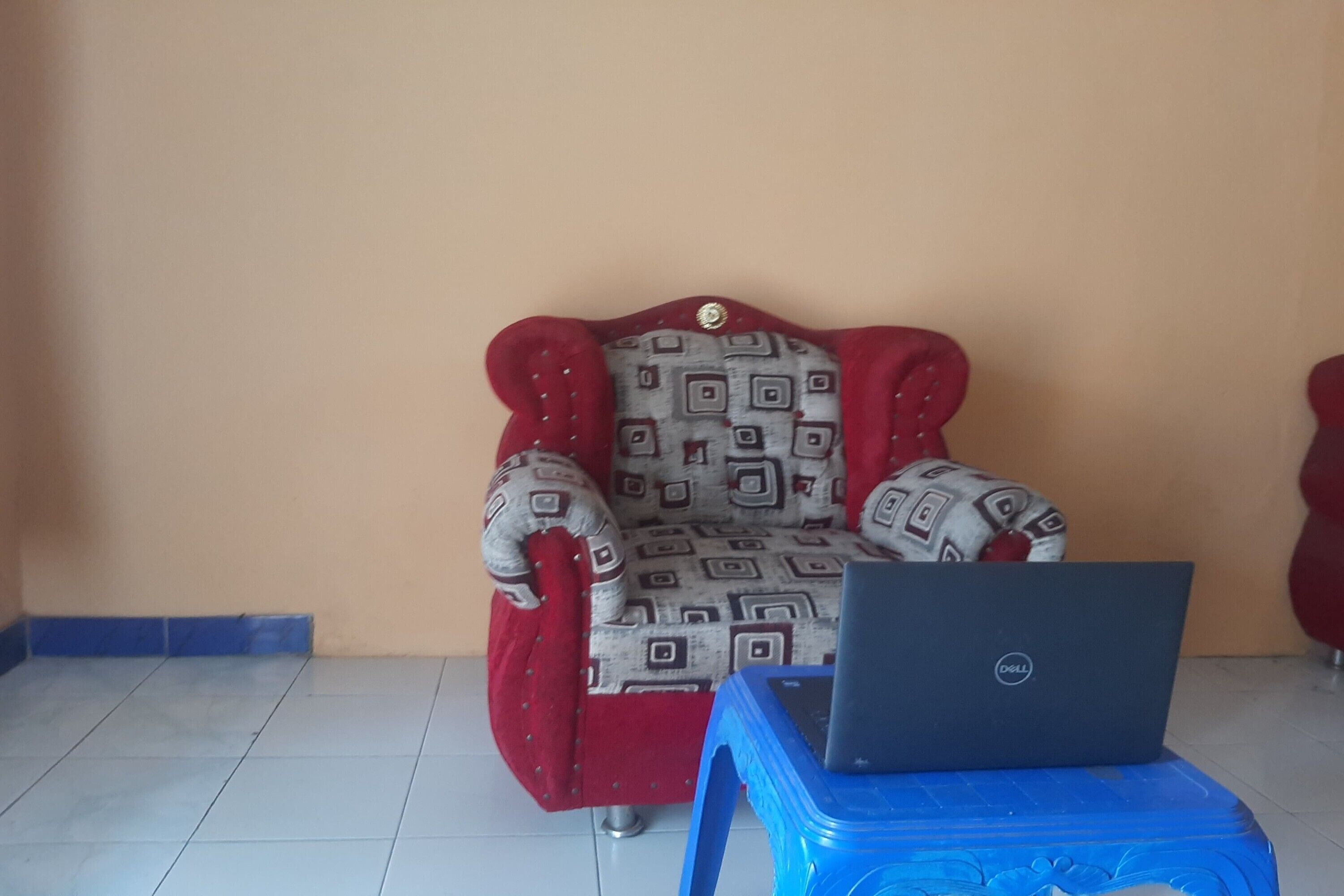 Uma poltrona vermelha com almofada estampada, uma mesa de plástico azul com um laptop e uma parede lisa.