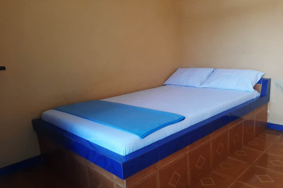 Ein Einzelbett mit blauem Kopfteil und weißer Bettwäsche in einem Zimmer mit schlichten Wänden und gemusterten Bodenfliesen.